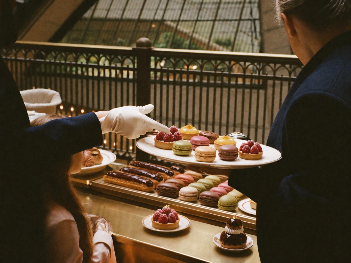 Une main gantée désigne une pâtisserie sur un plateau, sous la verrière d’un grand magasin parisien